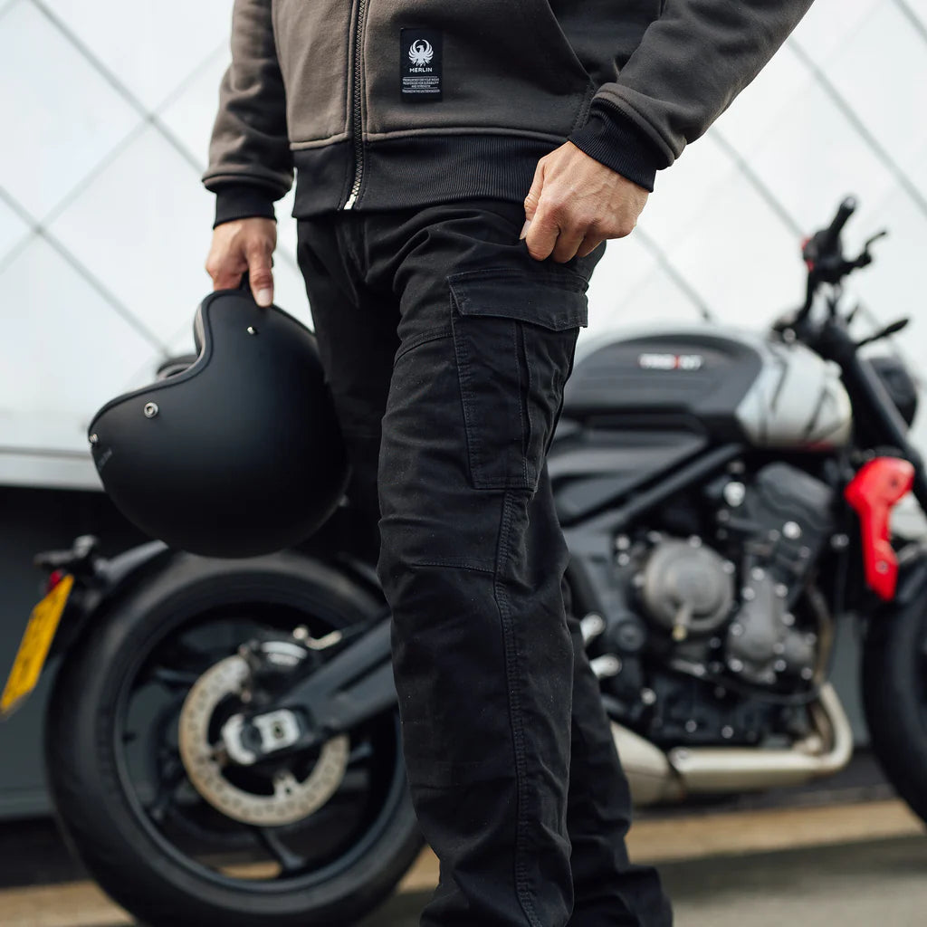 close up of Person wearing black Merlin Remy CE UKCA AA rated protective motorcycle trousers holding a merlin open face helmet with a blurred motorcycle background