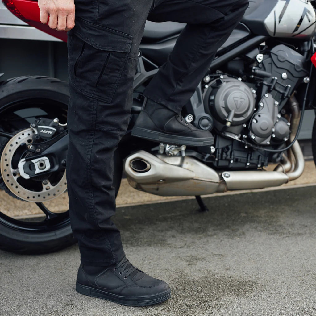 Person wearing black Merlin Remy protective motorcycle trousers standing next to a Triumph motorcycle in Cornwall.