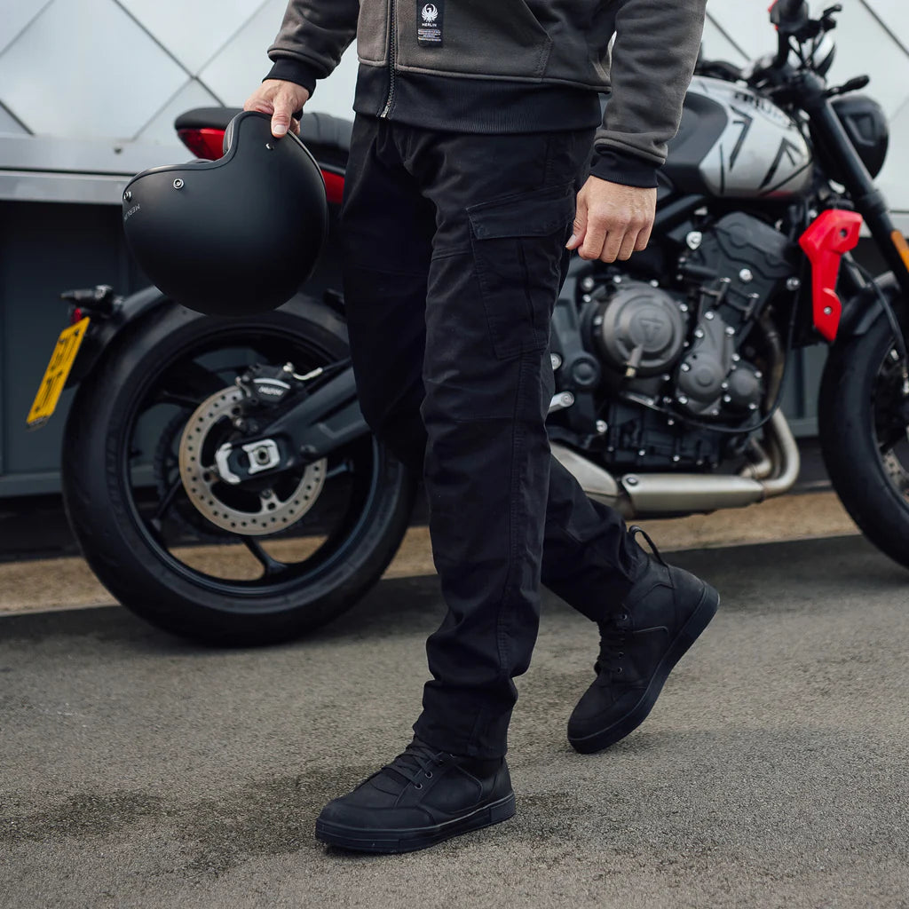 Person wearing black Merlin Remy CE UKCA AA rated protective motorcycle trousers holding a helmet with a motorcycle in the background