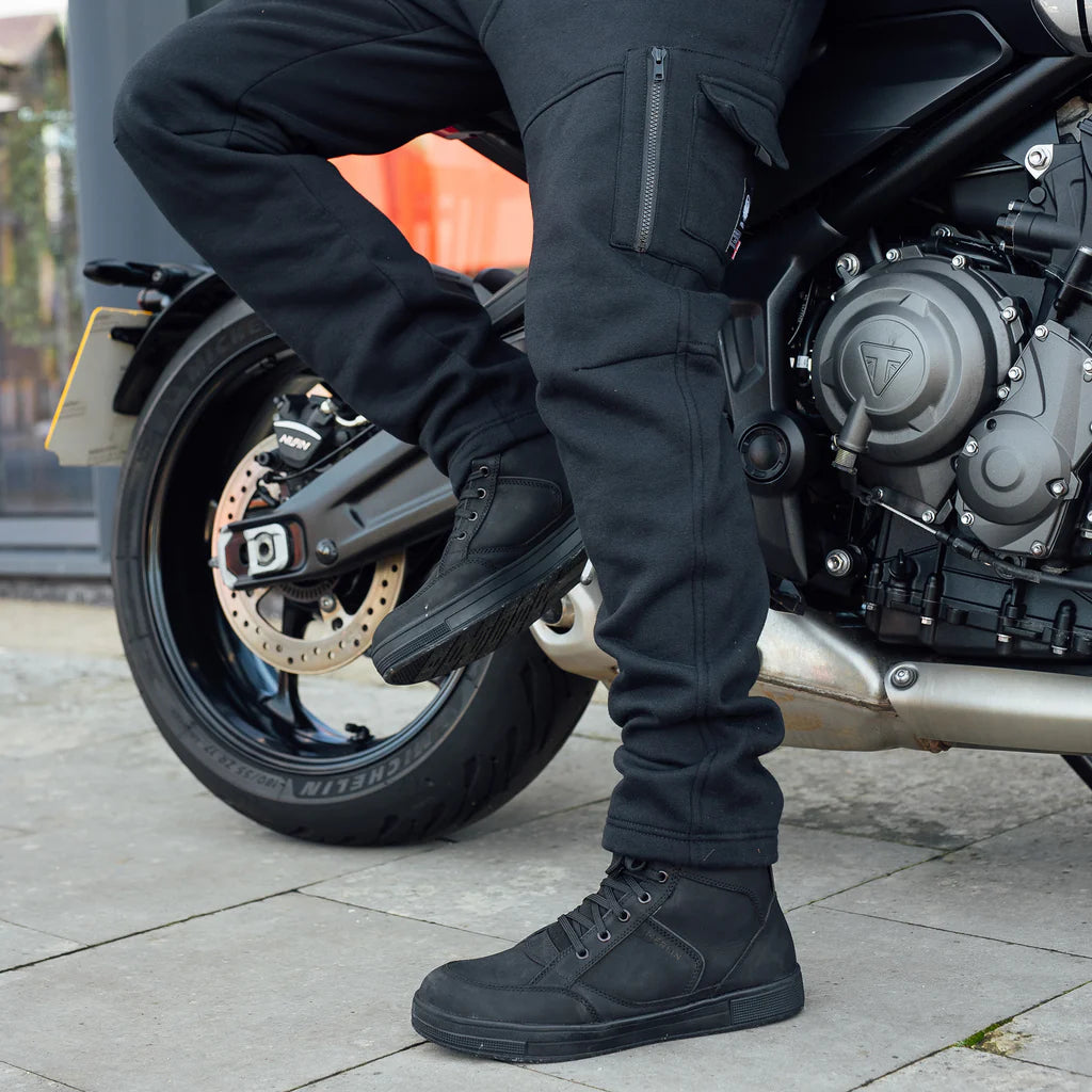 Person wearing black protective motorcycle Merlin Repton jogging bottoms standing next to Triumph motorcycle.
