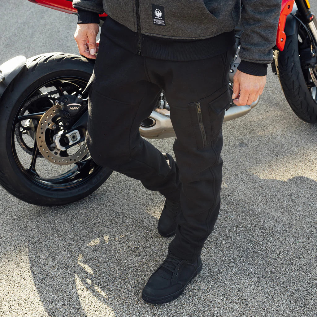 Person wearing black Merlin motorcycle joggers and protective trainers standing next to a motorcycle on a road.