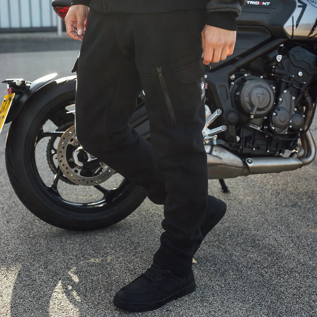 Person wearing black Merlin Repton CE AA rated protective jogger pants and shoes standing next to a Trident motorcycle on a road in Cornwall.