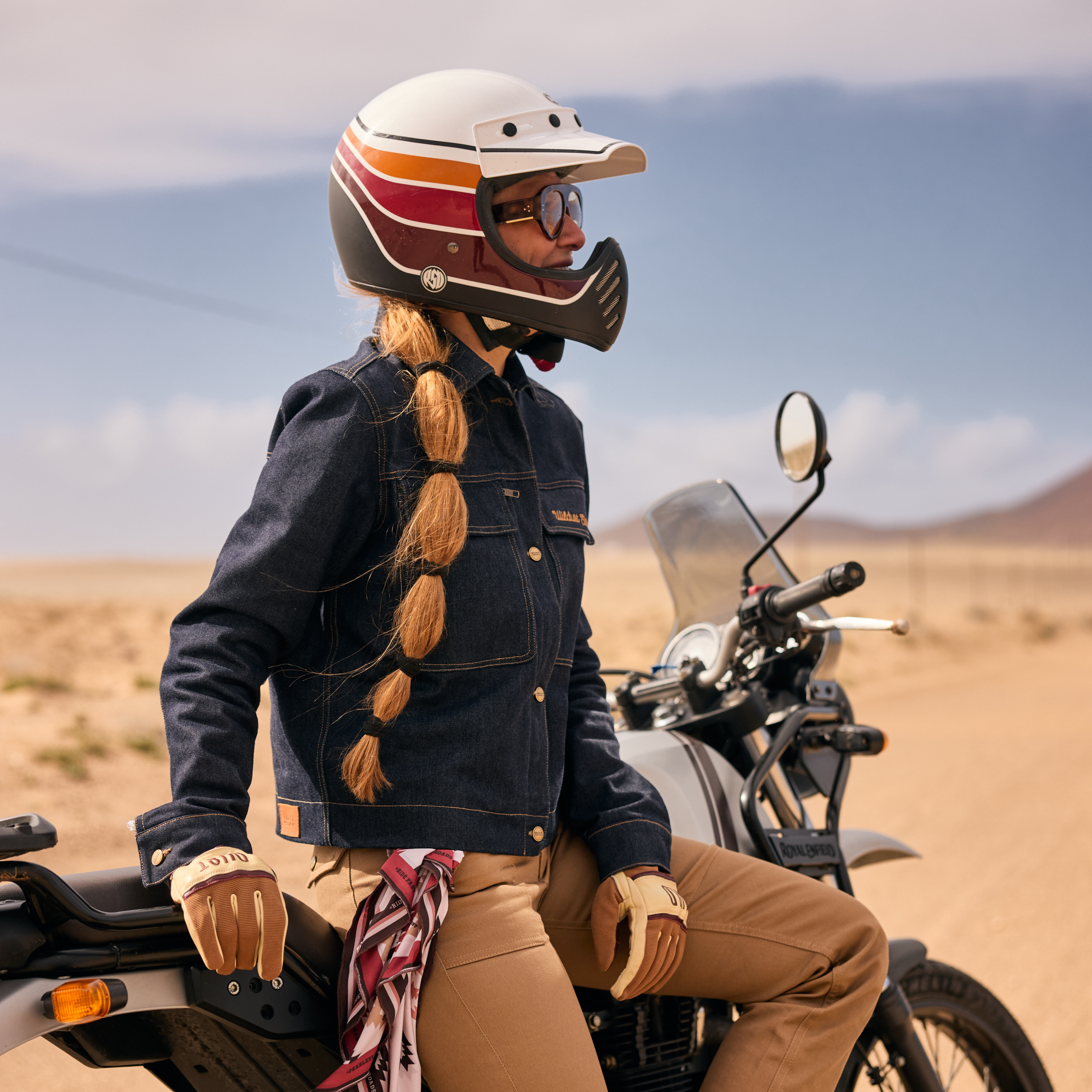 Person sitting on a royal enfield motorcycle in a desert landscape wearing a helmet and wildust sister heritage denim jacket.